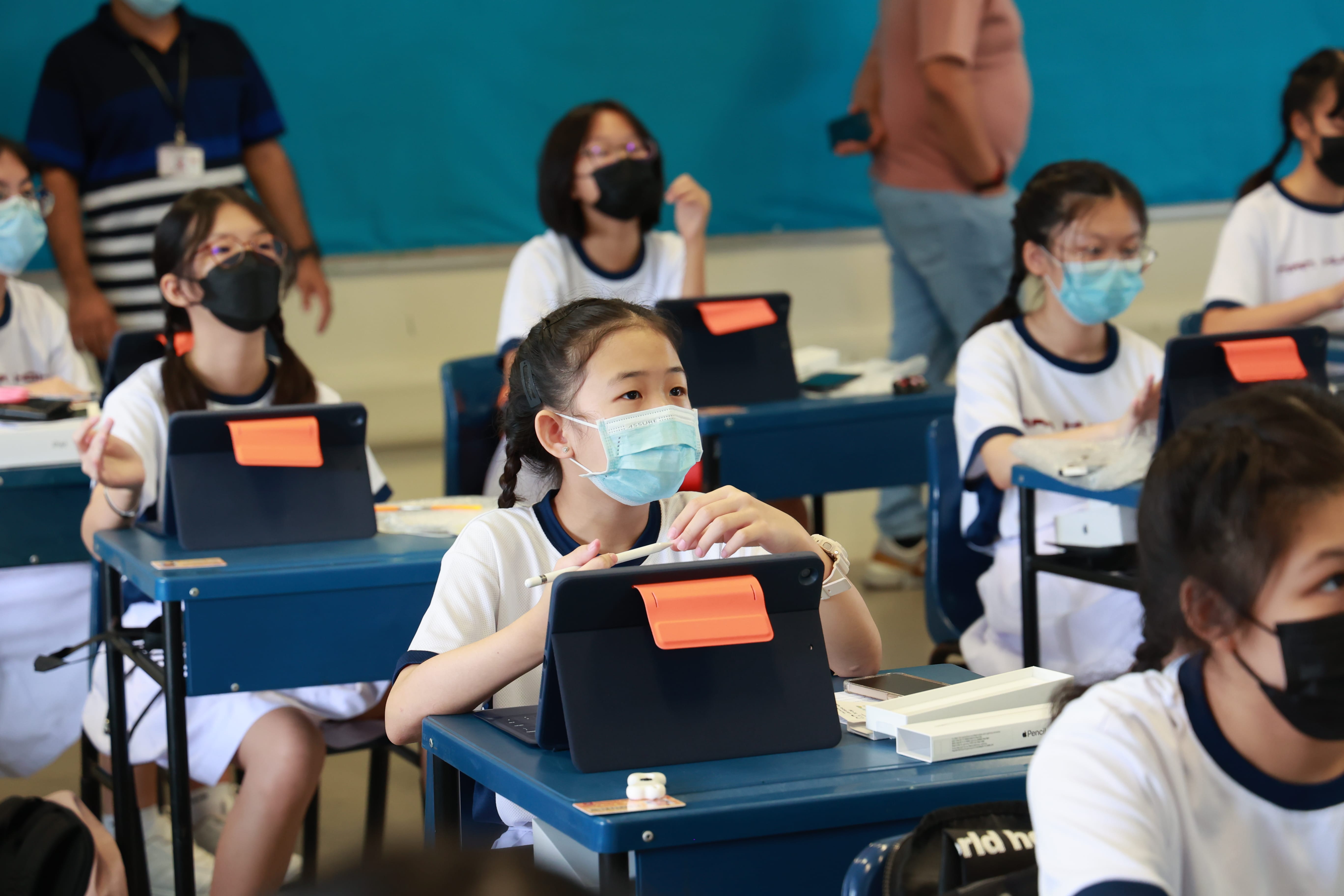 Students in masks sit at desks using tablets, with two adults standing in the back of the classroom.